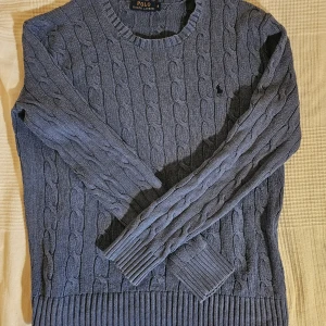 Blå kabelstickad tröja från Polo Ralph Lauren - Säljer en blå kabelstickad tröja från Polo Ralph Lauren. Tröjan har rund halsringning, långa ärmar och klassisk kabelstickad design. Perfekt för dig som gillar stilrena och tidlösa plagg.