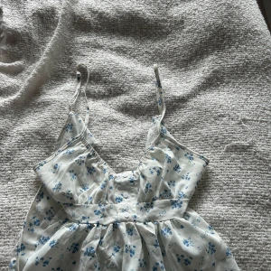 Blommig vit och blå linnetopp - Supersöt linnetopp med smala axelband och blommigt mönster i blått på vit botten. Toppen har knappar framtill och en stor knytrosett som detalj.