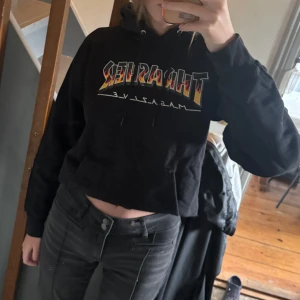 Svart Thrasher hoodie - Svart hoodie från Trasher. Lång modell (uppvikt på bilden) med snörning i huvan och magficka. Har ett mindre hål och trycket är något slitet, se bild 2 & 3. Priset kan diskuteras