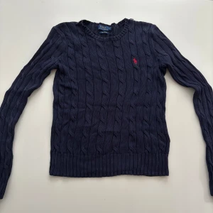 Kabelstickad tröja Polo Ralph Lauren - Två kabelstickade tröjor från Polo Ralph Lauren, en i mörkblått med röd broderad logga och en i ljusrosa med vit logga. Båda har rund hals, lång ärm och ribbade muddar. Klassisk och tidlös design i mjuk bomull, perfekt för lager-på-lager.