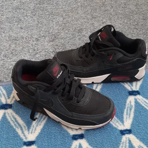 Nike Air Max svarta sneakers - Skor för barn. Svarta Nike Air Max sneakers med röda detaljer och vit sula. Klassisk chunky siluett med mesh och syntetmaterial, snörning och synlig Air Max-dämpning i sulan. Snygga kontraster mellan svart, vitt och rött gör dem till ett riktigt statement.