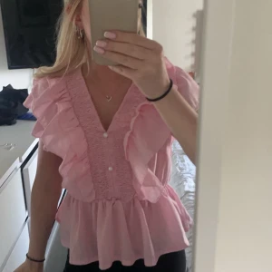 Rosa volangblus med v-ringning - Supersöt rosa blus med v-ringning och stora volanger längs axlar och bröst. Smockad framtill med små vita knappar och luftig, lätt material som ger en flowy känsla. Aldrig använt 