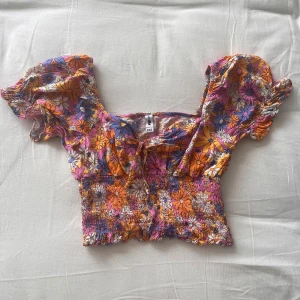 Blommig blus - Superfin croppad blus från NewYorker (FB Sister). Färgglatt blommönster i rosa, orange, lila, blått och vitt. Den har korta puffärmar, smockad midja och söt knytning framtill. Skärningen vid bysten gör att den sitter väldigt fint. Perfekt nu till sommaren🌸💐 
