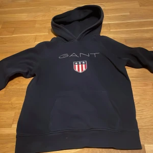 Svart hoodie från GANT med logga - Svart hoodie från GANT med broderad logga och sköld på bröstet. Klassisk känguruficka framtill och stor huva. Mjuk och skön, perfekt för chill dagar. Långärmad modell i bomullsmix. Skick 9/10