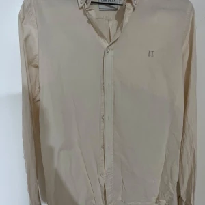 Beige skjorta från Les Deux, strl S - Stilren beige skjorta från Les Deux med klassisk krage och knäppning framtill. Skjortan har lång ärm och diskret broderad logga 'II' på bröstet. Perfekt för dig som gillar enkel och clean stil.