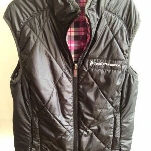 Quiltad snygg dunväst från Peak Performance - Snygg quiltad dunväst från Peak Performance med svart utsida och dragkedja. Insidan är fodrad i ett färgstarkt rutigt mönster i rosa, vit och svart. Västen har hög krage och är tillverkad i polyester. Perfekt för lager-på-lager och sportig stil. Vändbar modell.