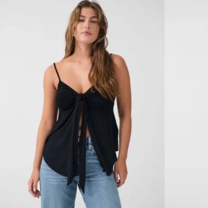 Svart linnetopp med knyt framtill - Svart linnetopp med smala axelband och snygg knytning framtill. Toppen har en lös passform och djup v-ringning, perfekt för varma dagar. Tunt och mjukt material som ger en avslappnad vibe.
