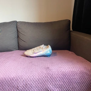 Nike Mercurial - Säljer ett par Nike fotbollsskor i vitt med snygga blå och lila detaljer. Skorna har snörning och en cool, skimrande sula i blått och lila. Perfekta för dig som vill sticka ut på planen med stilrena och moderna skor. Nypris ligger på 3500kr