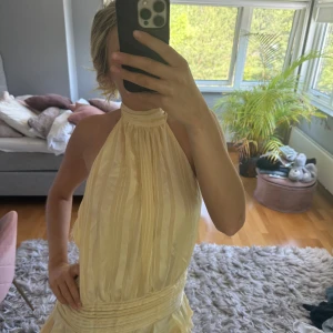 Pepparmayo gul klänning - Säljer en champagnefärgad omlottklänning från  med halterneck och knyt i nacken. Prislapp kvar då jag beställde fel storlek. Klänningen har subtila ränder, markerad midja och volangdetalj nedtill. Öppen rygg och lätt glansigt material ger en lyxig känsla.