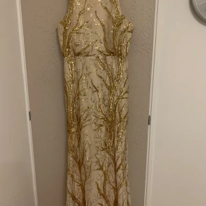 Långklänning med guldpaljetter - Superfin långklänning i ljus beige med guldiga paljetter i ett grenliknande mönster. Klänningen är ärmlös med smala axelband och har dragkedja i ryggen. Perfekt för bal eller nyår, ger en riktigt lyxig och glittrig känsla. Använt 1 gång. Sitter jättefint på kroppen. Köpte på Whoops för 1500kr. Stl XL, men passar M- L. 