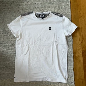Vit Weekend Offender t-shirt med rutiga detaljer - Vit t-shirt från Weekend Offender med rutiga detaljer i svart, grått och beige vid axlar och insida krage. Klassisk passform och liten svart logopatch på bröstet. Tillverkad i mjuk bomull, perfekt för en clean och stilren look.