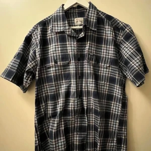 Rutig kortärmad skjorta - Snygg kortärmad skjorta i mörkblått och vitt rutigt mönster. Skjortan har klassisk krage, två bröstfickor med lock och knappar framtill. Perfekt för dig som gillar en avslappnad men ändå stilren look.