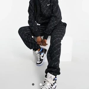 Svart Nike half zip hoodie och byxor med mönster - Svart tracksuit från Nike med half zip och helöverdraget mönster av grå Nike Swoosh-loggor. Tröjan har hög krage, långa ärmar och ribbade muddar. Perfekt för dig som gillar sportig och street-inspirerad stil.