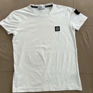 Vit Stone Island t-shirt med patch - Vit t-shirt från Stone Island i bomull med klassisk rund hals och korta ärmar. Ikonisk svart och gul Stone Island patch sitter på vänster ärm och en liten logotyp på bröstet. Enkel och clean design som passar till allt.