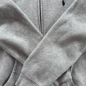 Grå hoodie från Polo Ralph Lauren - Klassisk grå hoodie från Polo Ralph Lauren med dragkedja framtill och broderad marinblå logga på bröstet. Tröjan har huva, känguruficka och är tillverkad i mjuk bomullsmix. Perfekt för chill dagar eller när du vill ha en enkel och stilren look.