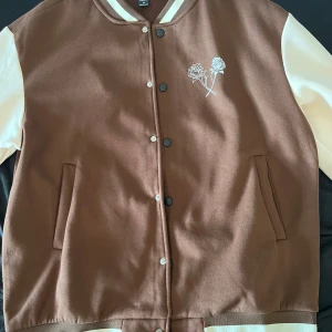 Brun och vit varsityjacka med rosdetalj - Säljer en brun och vit varsityjacka från Tworopes med tryckta rosor på bröstet och logga på ärmen. Jackan har knappar framtill, två fickor och kontrastfärgade ärmar. Perfekt för dig som gillar streetwear och snygga detaljer.