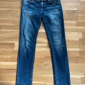 Blå jeans från Nudie Jeans - Säljer ett par klassiska blå jeans från Nudie Jeans med orangea sömmar och fyra fickor. Jeansen har en rak passform och subtila slitningar framtill och baktill. Perfekta för dig som gillar en tidlös denimlook.