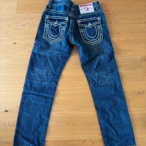Blå jeans True Religion - Säljer ett par klassiska blå jeans från True Religion med tydliga vita kontrastsömmar och stora bakfickor med lock och knapp. Byxorna har rak passform och är tillverkade i denim. Märkeslapp bak i midjan.