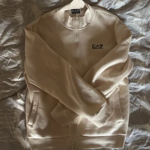 Beige ziptröja från Emporio Armani EA7 - Säljer en stilren beige ziptröja från Emporio Armani EA7. Jackan har dragkedja framtill, två fickor och EA7-logga på bröstet. Perfekt för en avslappnad och snygg look. Endast använd två gånger.Säljs pga för liten