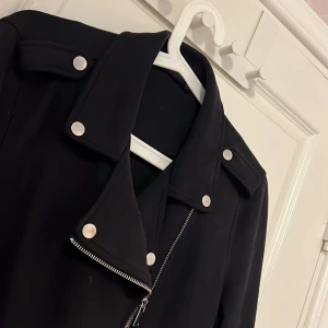 Svart bikerjacka från KappAhl - Säljer en svart bikerjacka från KappAhl med silvriga dragkedjor, knappar och bälte. Jackan har klassisk krage och detaljer som ger en cool look. Perfekt för dig som vill ha en stilren och edgy jacka. Storlek M 
