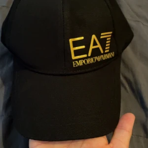 Ea7 keps  - Säljer en snygg och stilren keps från Emporio Armani EA7. Svart bas med exklusiv guldig logga framtill. Fick den som present men använder inte kepsar, så den är helt oanvänd och i nyskick. Passar både herr och dam. Justerbar baktill för perfekt passform