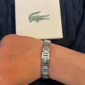 Lacoste armband  - Hej säljer min Lacoste armband Bara testat ett par gånger, fortfarande i nyskick.Kommer med originalask och påse.Snygg, stilren modell med Lacoste-logga – perfekt till både vardag och fest. Köpt  sommaren 2024