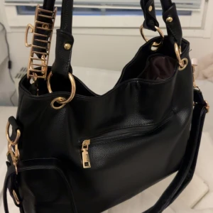 Svart väska - Säljer denna snygga och praktiska vintage väskan, svart med guld detaljer!! Perfekt att ha skoldatorn och andra saker i 🌟🌟 Aldrig använd, endast provat att ha saker i, därför helt ny (köpt för någon vecka sedan). Den har massa fickor och dragkedjor inuti också, skriv för mer detaljer isåfall 💕Kommer med ett långt band (andra bilden) och handtag (första bilden), handtagen kan man ha på axeln också. Hårklämman och den lilla rosa rosetten följs inte med!!