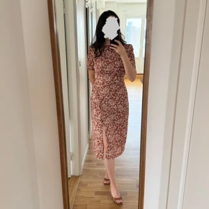 Blommig klänning med slits - Säljer en rödbrun klänning med vitt blommönster och korta ärmar. Klänningen har hög hals, figurnära passform och en snygg slits framtill. Perfekt för vår och sommar! Materialet känns lätt och bekvämt. På lappen står det strl 40 men denna sitter som en storlek 38, så köp den med rätt förväntan.
