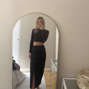Svart långkjol med slits - Säljer en svart långkjol med hög midja och en snygg slits framtill. Kjolen har en stilren och enkel design som passar perfekt till många olika looks. Materialet känns mjukt och följsamt.
