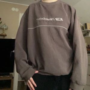 Gråbrun sweatshirt från Reebok - En gråbrun sweatshirt från Reebok med broderad logga och texten 'Authentic' på bröstet. Tröjan har rund halsringning och kontrastpaneler i ljusgrått på sidorna. Perfekt för en sportig och avslappnad stil.