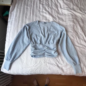 Ljusblå omlottopp från Gina Tricot - Supersöt ljusblå omlottopp från Gina Tricot i smockat tyg. Toppen har lång ärm med breda muddar, v-ringning och croppad passform. Perfekt att styla med höga jeans eller kjol för en chill men trendig look.