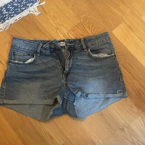 Blå jeansshorts med rå kant - Snygga blå jeansshorts från Gina Tricot med klassisk femficksdesign och råa kanter nertill. Shortsen har normal passform, bälteshällor och dragkedja med knapp. Perfekta för sommardagar och enkla att matcha med allt.