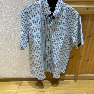 Rutig kortärmad skjorta - Kortärmad skjorta med klassiskt rutmönster i blått, grönt och rött på vit bas. Skjortan har knappar framtill och en bröstficka. Perfekt för varma dagar och enkel att matcha med jeans eller shorts.