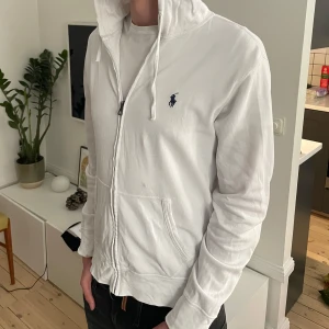 Vit hoodie från Polo Ralph Lauren - Vit hoodie från Polo Ralph Lauren med dragkedja, snörning i huvan och klassisk blå logga på bröstet. Tillverkad i mjuk bomull, har känguruficka och långa ärmar. Perfekt för en clean och stilren look. OBS den har några fläckar t.ex den som syns på bild 1!