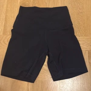 Svarta träningstights-shorts Lululemon - Svarta cykelshorts från Lululemon med hög midja och tight passform. Tillverkade i ett stretchigt och mjukt syntetmaterial som passar perfekt för träning. Klassisk design med diskret logga bak på linningen.