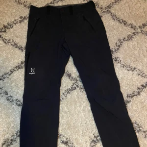 Svarta softshellbyxor från Haglöfs - Svarta softshellbyxor från Haglöfs med dragkedjeförsedda fickor och diskret logga på låret. Byxorna är i ett slitstarkt syntetmaterial och har en rak passform som funkar perfekt för utomhusaktiviteter. Snygga detaljer och bekväm midja.