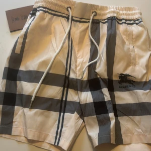 Burberry rutig shorts med snörning - Snygga Burberry shorts i klassiskt beige med svart och grått rutmönster. De har elastisk midja med vit snörning och sidofickor. Perfekta för sommaren och tillverkade i ett lätt syntetmaterial. Säljer pga försmå. Sitter mer som m än L storleksvis. Pris kan sänkas vid snabb affär