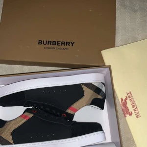 Burberry skor - Snygga svarta sneakers från Burberry med klassiskt rutigt mönster i beige, svart och rött på hälen. Skorna har vita platta sulor, svarta snören och röda detaljer vid snörningen. Perfekt för dig som vill ha en stilren men ändå unik look. Present från min vän men för stora för mig HELT NU PASSAR OCKSÅ STORLEK 42