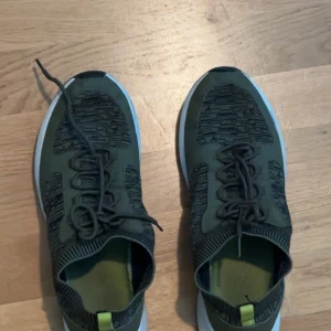 Olivgröna sneakers med stickade detaljer - Säljer ett par olivgröna sneakers med stickade detaljer och vit sula. Skorna har snörning och en sportig design med inslag av svart mönster på ovandelen. Perfekta för dig som gillar en modern och bekväm stil.