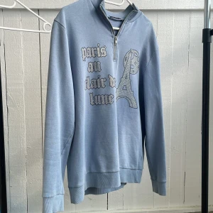 Ljusblå hoodie med dragkedja och Paris-tryck - Säljer en blå hoodie/sweatshirt som är i helt nytt skick, använd ungefär 5 gånger och säljes pågrund av att jag inte använder den.
