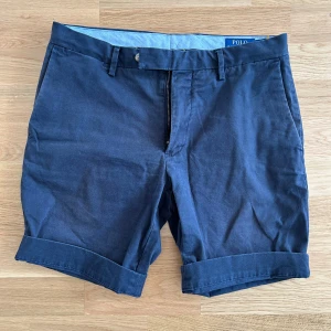 Mörkblå shorts från Polo Ralph Lauren - Snygga mörkblå shorts från Polo Ralph Lauren med klassisk passform. Shortsen har uppvikta benslut, bälteshällor, dragkedja och knäppning fram samt två sidofickor och två bakfickor med knapp.