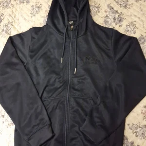 Svart tröja från Hugo Boss - Säljer en hoodie från Hugo Boss med dragkedja och huva. Tröjan har snörning i huvan och två fickor framtill. Perfekt för en avslappnad stil och enkel att matcha med det mesta.