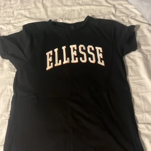 Svart t-shirt från Ellesse - Säljer en svart t-shirt från Ellesse med stor vit och orange logga på bröstet. T-shirten har rund halsringning och korta ärmar. Perfekt för en sportig och avslappnad stil. Aldrig använd.