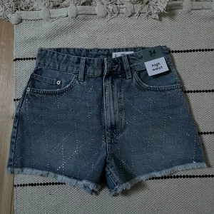 Glittriga jeansshorts från Lefties - Snygga ljusblå jeansshorts från Lefties med hög midja och råa fransade kanter. Shortsen har glittriga detaljer framtill som ger extra edge och klassisk femficksmodell. Perfekta för sommardagar när du vill sticka ut lite extra.