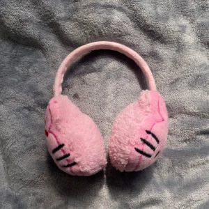 Rosa fluffiga Hello Kitty öronvärmare - Supergulliga rosa öronvärmare med Hello Kitty-design, broderade detaljer och en liten röd blomma på ena sidan. Mjukt och fluffigt material som håller dig varm och söt under kalla dagar. Perfekt accessoar för dig som älskar kawaii-stil!