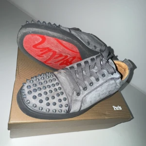 Gråa louboutins - Säljer ett par gråa sneakers från Christian Louboutin med svarta nitar på tån och sidorna. Skorna har klassisk röd sula, snörning och är tillverkade i mocka. Perfekta för dig som vill sticka ut med en edgy stil. Dynpåsen kommer med också