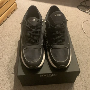 Svarta sneakers från Mallet med reflexdetaljer - Säljer ett par svarta Mallet sneakers med vita sulor och snygga reflexdetaljer som lyser upp i blixtljus. Skorna har snörning och diskret camouflagemönster på ovandelen. Perfekta för dig som vill sticka ut med stilrena detaljer.