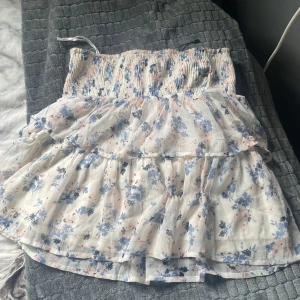 Blommig volangklänning från Vero Moda - Supersöt kortklänning från Vero Moda med blommigt mönster i blått och rosa på vit botten. Klänningen är ärmlös, har smock upptill och volanger i kjolen för en luftig känsla. Perfekt för sommarens alla tillfällen!