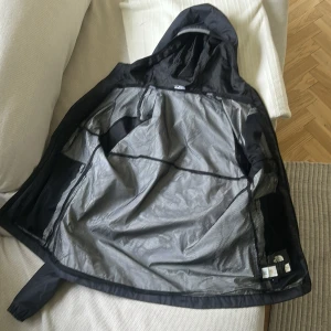 Svart vindjacka från The North Face - Säljer en svart vindjacka från The North Face med huva och dragkedja framtill. Jackan har två sidofickor med dragkedja och är tillverkad i ett lätt syntetmaterial. Perfekt för blåsiga dagar och enkel att packa ner i väskan.