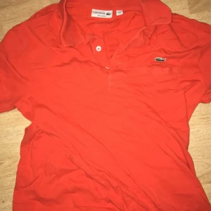 Röd pikétröja från Lacoste - Säljer en klassisk röd pikétröja från Lacoste med korta ärmar och den ikoniska krokodilloggan på bröstet. Perfekt för en sportig och avslappnad stil.